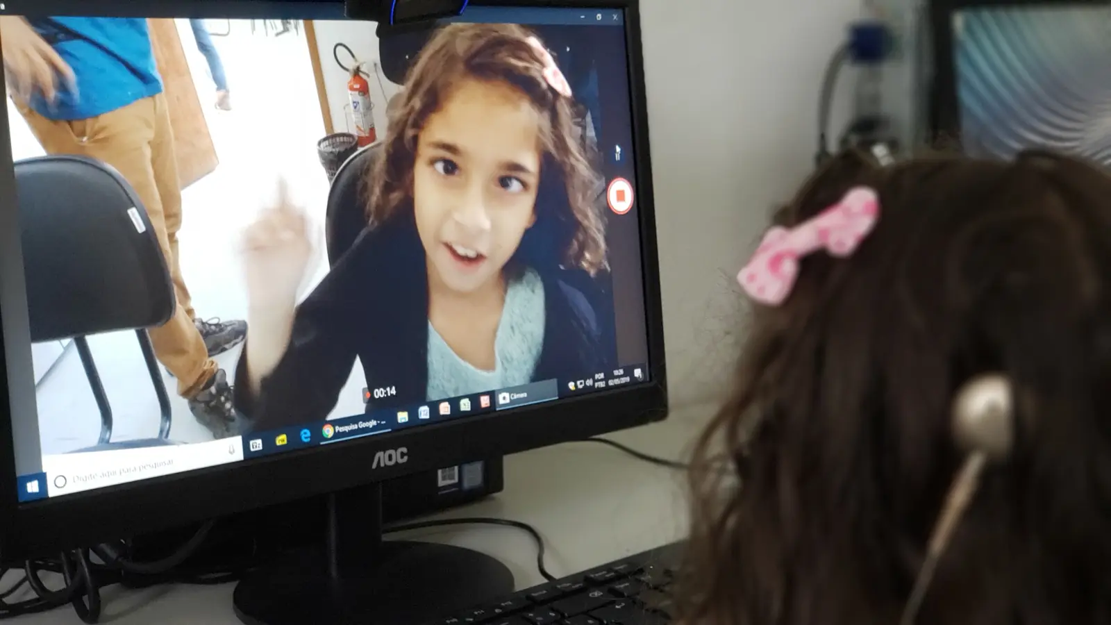 Menina conversando em Libras por videochamada, vista por trás de outra criança com aparelho auditivo